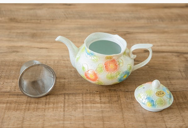 Decorative ceramic teapot with floral patterns on a wooden surface, accompanied by a metal tea strainer and lid.