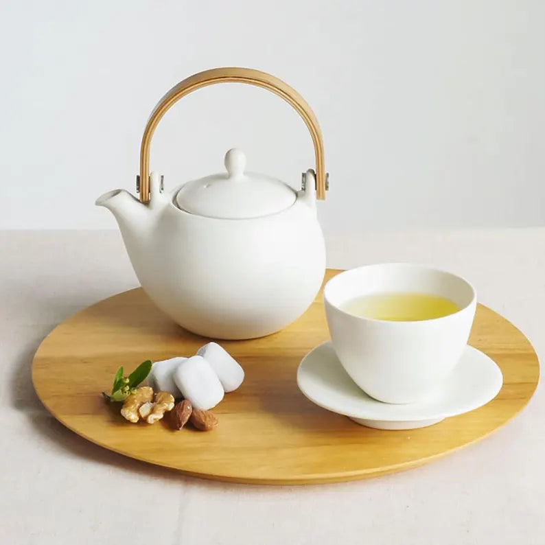 White teapot and cup with wooden handle on a wooden tray with tea leaves and nuts.