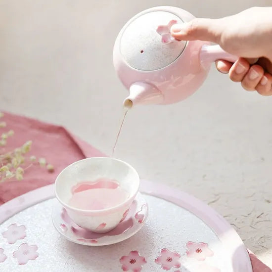 Person pouring tea from a pink teapot into a matching teacup on a white surface.