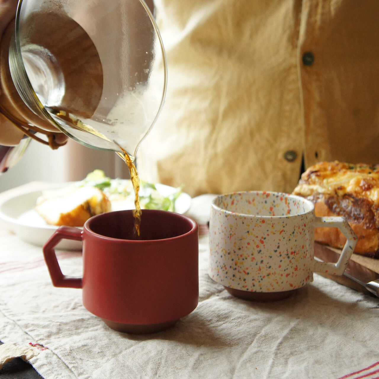 Person pouring coffee from a carafe into a red mug with a speckled white mug on a table.