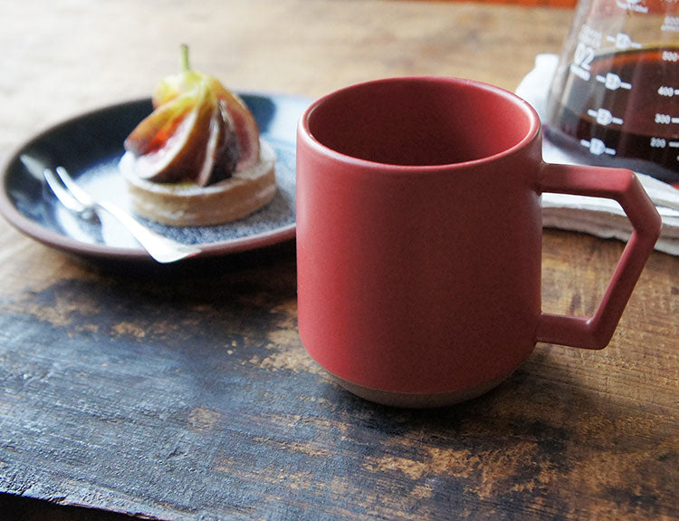 Red mug on a wooden table with a plate of food and a carafe in the background. Japanese Ceramic. Made in Japan