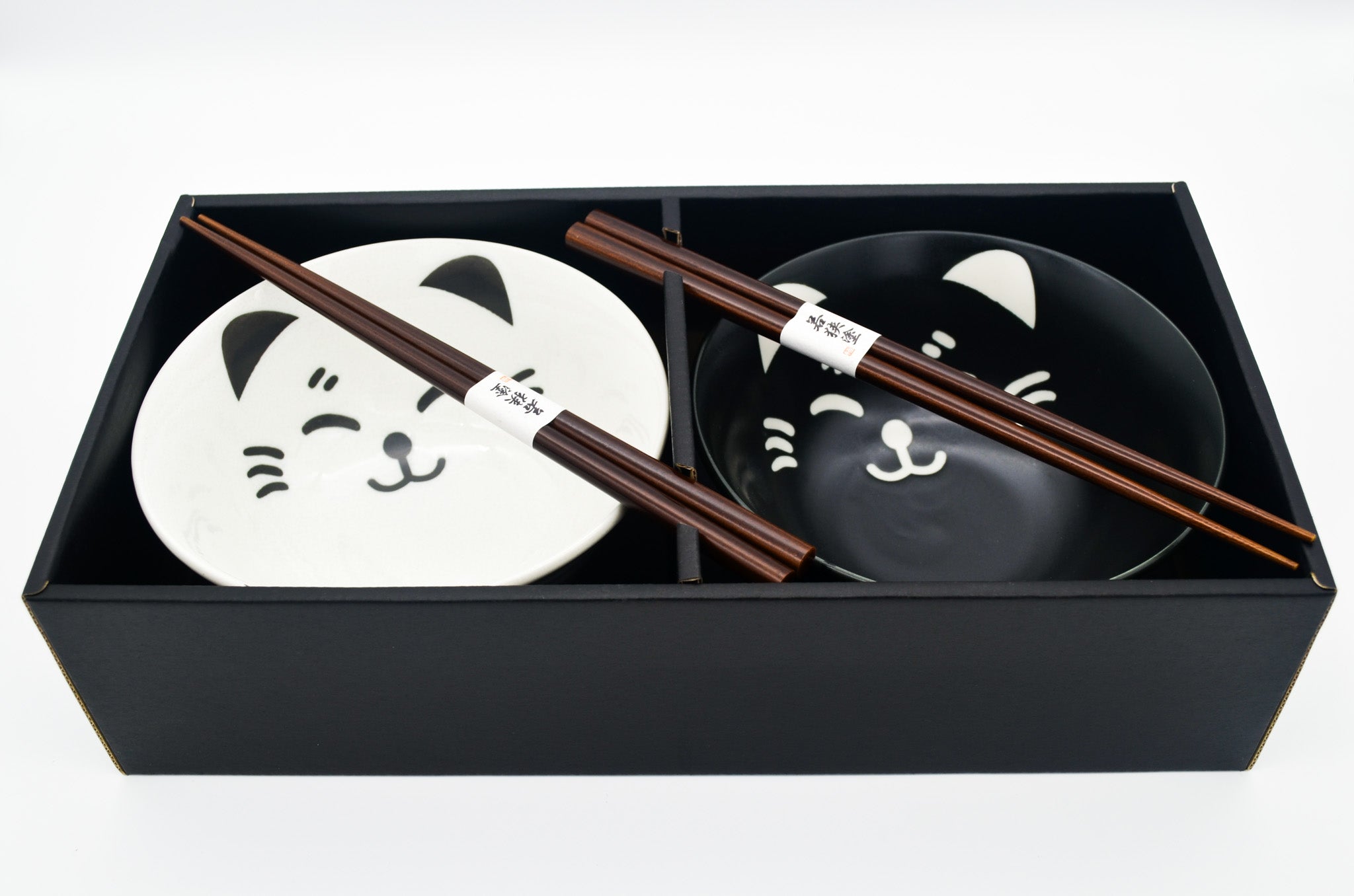 Two black bowls with cat face designs and wooden chopsticks in a black box on a white background
