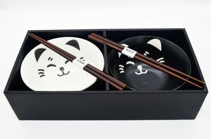 Two black bowls with cat face designs and wooden chopsticks in a black box on a white background
