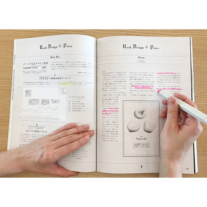 Open book with text and diagrams on a wooden surface, held by a hand with a pen.