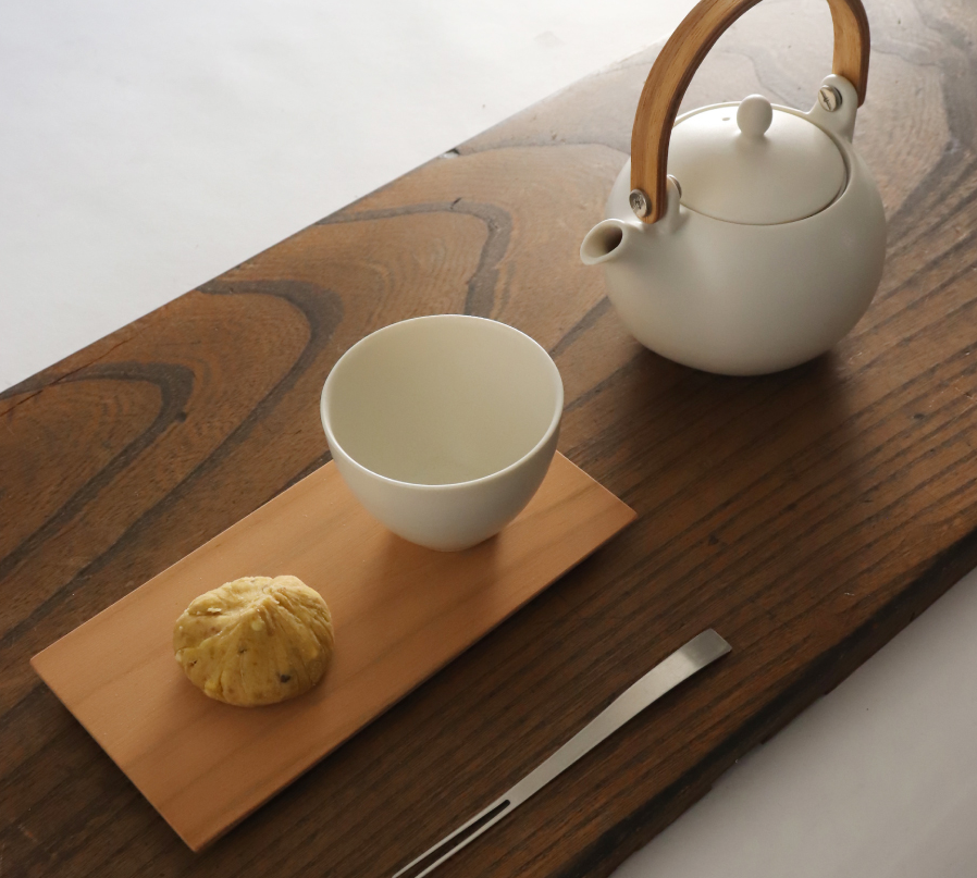 White teapot and cup on a wooden tray with a wooden object and metal utensil.