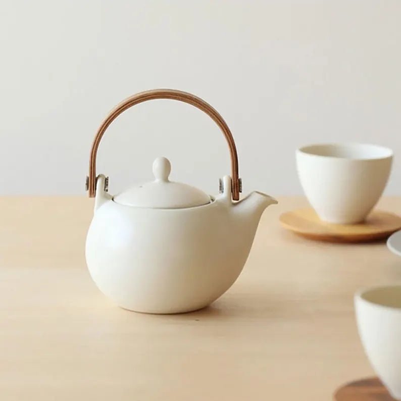 White ceramic teapot with wooden handle and lid on a beige surface.